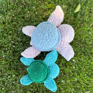May include: Two crocheted turtle toys. The larger turtle is light blue and pink, while the smaller turtle is green and blue.