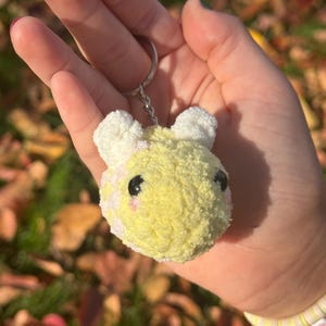 May include: A fluffy yellow and white bear head keychain. The bear has black eyes and pink cheeks. The keychain is attached to a silver keyring. The plush toy is made of soft material.