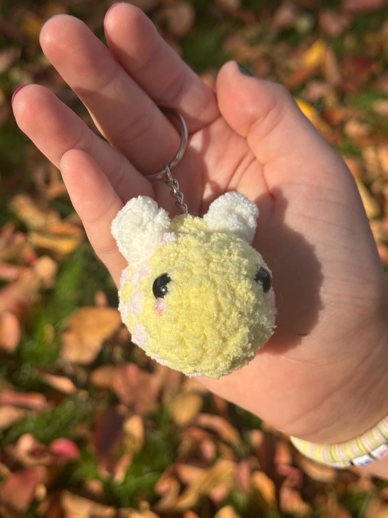 May include: A fluffy yellow and white bear head keychain. The bear has black eyes and pink cheeks. The keychain is attached to a silver keyring. The plush toy is made of soft material.