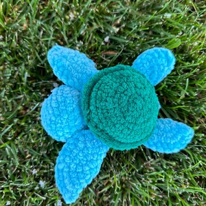 May include: A plush toy turtle with a blue shell and a green head. The turtle is lying on its back on a bed of green grass.
