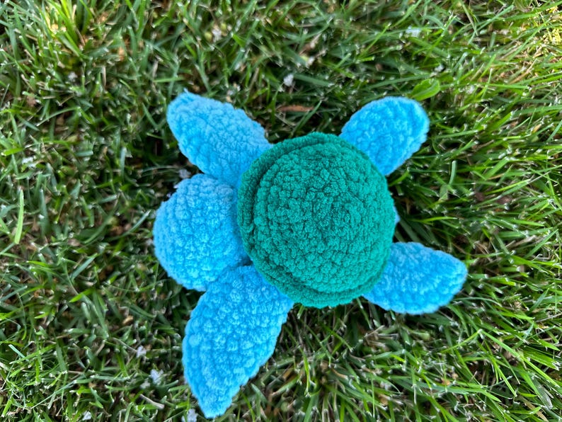 May include: A plush toy turtle with a blue shell and a green head. The turtle is lying on its back on a bed of green grass.