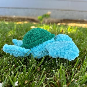 May include: A crocheted turtle toy with a green shell and blue body. The toy is lying on a bed of green grass.