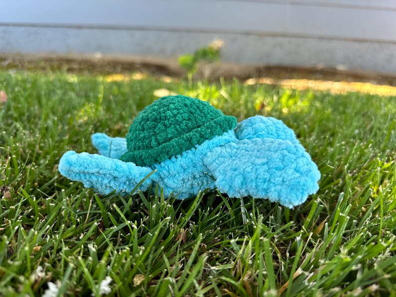May include: A crocheted turtle toy with a green shell and blue body. The toy is lying on a bed of green grass.