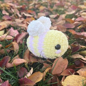 May include: A soft, handmade bee toy with yellow and lilac stripes, black eyes, and white wings. The bee is set amongst green grass and fallen autumn leaves.