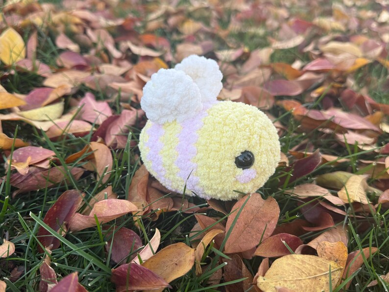 May include: A soft, handmade bee toy with yellow and lilac stripes, black eyes, and white wings. The bee is set amongst green grass and fallen autumn leaves.