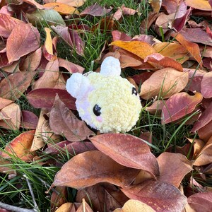 May include: A handmade, yellow and white plush bee keyring with black eyes and pink cheeks. The bee is surrounded by fallen autumn leaves in shades of red, brown, and orange, with patches of green grass.