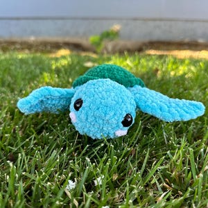 May include: A crocheted blue and green turtle toy with black eyes and a white nose. The toy is sitting in green grass.