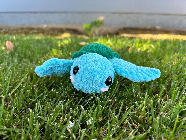 May include: A crocheted blue and green turtle toy with black eyes and a white nose. The toy is sitting in green grass.