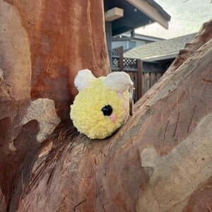 May include: A yellow and white plush bee keyring with a pink cheek and black eye. The bee is round and fluffy, with a small metal ring attached. The keyring is set against a tree trunk.