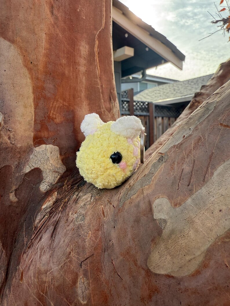 May include: A yellow and white plush bee keyring with a pink cheek and black eye. The bee is round and fluffy, with a small metal ring attached. The keyring is set against a tree trunk.