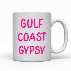 May include: White ceramic mug with the words "GULF COAST GYPSY" in bright pink, bold, rounded letters. The mug has a standard handle and is set against a plain white background. A simple, everyday coffee mug.
