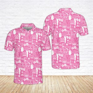 May include: Pink golf polo shirt with a white pattern of golf-related phrases and images. The shirt features a collared neckline and short sleeves. The design includes golf carts, golf clubs, and text like "Swing" and "Warm Up".
