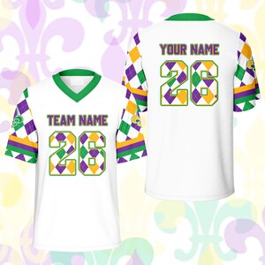 Op de afbeelding: Wit shirt met groene, paarse en gouden accenten. Het shirt heeft het nummer 26 in een ruitpatroon, met de woorden "TEAM NAME" en "YOUR NAME" op de voor- en achterkant. De mouwen hebben een ruitpatroon.