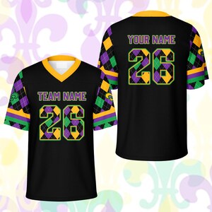 Op de afbeelding: Zwart voetbalshirt met gouden, paarse en groene accenten. De voorkant toont "TEAM NAME" en het nummer "26" in een ruitpatroon. De achterkant heeft "YOUR NAME" en het nummer "26".