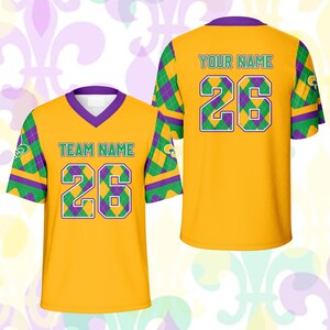 Op de afbeelding: Twee gele voetbalshirts met paarse en groene accenten. Het shirt aan de voorkant heeft "TEAM NAME" en het nummer "26" in groen. Het shirt aan de achterkant heeft "YOUR NAME" en het nummer "26". De mouwen hebben een ruitpatroon.