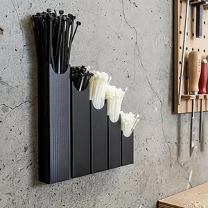 May include: A black cable tie organiser mounted on a concrete wall. The organiser has five compartments, each holding a different quantity of black and white cable ties. A pegboard with tools is visible in the background.
