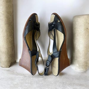 May include: A pair of black leather wedge sandals featuring a knotted front strap, an adjustable ankle strap with a metal buckle, and a wood-grain textured wedge heel. The interior footbed is light beige with the Clarks brand logo visible.