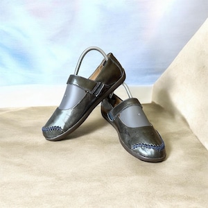 May include: A pair of dark olive green flats with a glossy finish, featuring a single strap across the instep with a hook-and-loop closure and decorative dark blue cross-stitching detail along the rounded toe area of each shoe.