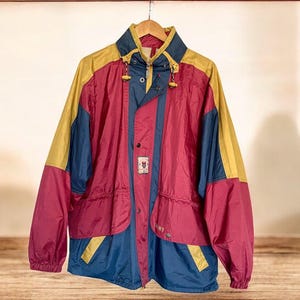 以下が含まれることがあります： 赤、黄色、紺色の色使いが特徴的なヴィンテージジャケット。高い襟、ジッパー、スナップ留めが特徴です。フロントポケットが2つあり、胸元には小さなエンブレムが付いています。木製のハンガーに掛けられています。