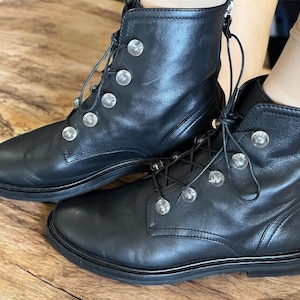 Könnte beinhalten: Ein Paar schwarze Leder-Ankle-Boots mit silberfarbenen, kreisförmigen Verzierungen. Die Stiefel haben ein Schnürdesign und eine runde Spitze. Die Stiefel werden auf einer Holzoberfläche präsentiert.