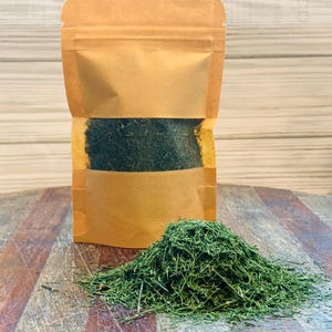 May include: A kraft paper bag filled with dried green herbs, with a small pile of the herbs in front of the bag. The bag is sealed at the top and has a clear window to see the contents. The herbs are a vibrant green color.