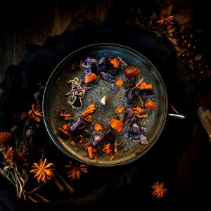 May include: A round black candle in a container, topped with silver glitter, orange and purple dried flower petals, small polished stones, and a small metal charm of a black cat wearing a purple witch hat, featuring a single central wick.