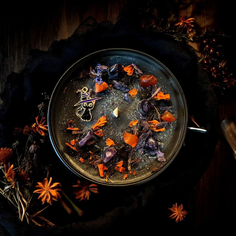 May include: A round black candle in a container, topped with silver glitter, orange and purple dried flower petals, small polished stones, and a small metal charm of a black cat wearing a purple witch hat, featuring a single central wick.