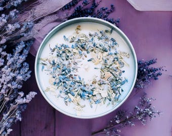 May include: A round candle in a light blue metal tin, filled with white wax and topped with a circular arrangement of dried blue lavender buds and small light-colored botanical pieces, centered around a single white cotton candle wick.