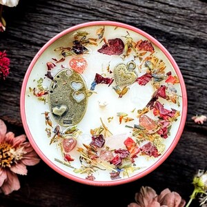 May include: A round candle in a pink container featuring a white wax surface topped with dried flower petals, small pink crystals, and two metallic charms, one oval-shaped with heart cutouts and one small heart-shaped charm with a textured surface.