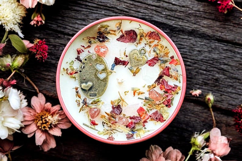 May include: A round candle in a pink container featuring a white wax surface topped with dried flower petals, small pink crystals, and two metallic charms, one oval-shaped with heart cutouts and one small heart-shaped charm with a textured surface.