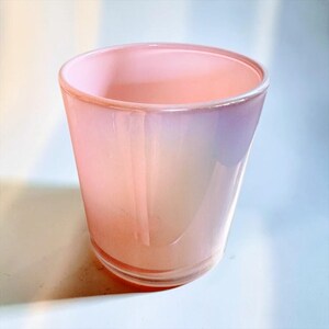 May include: A cylindrical glass vessel featuring a soft, pale pink hue with a subtle iridescent finish. The container has a smooth, polished surface and a clear, thick base, presenting a simple, open-top design with a uniform, slightly tapered shape.