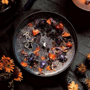 May include: A round black candle in a metal tin, topped with dark wax, silver glitter, dried orange flower petals, purple crystal chips, and a small metal charm of a black cat wearing a purple witch hat, centered around a single white wick.