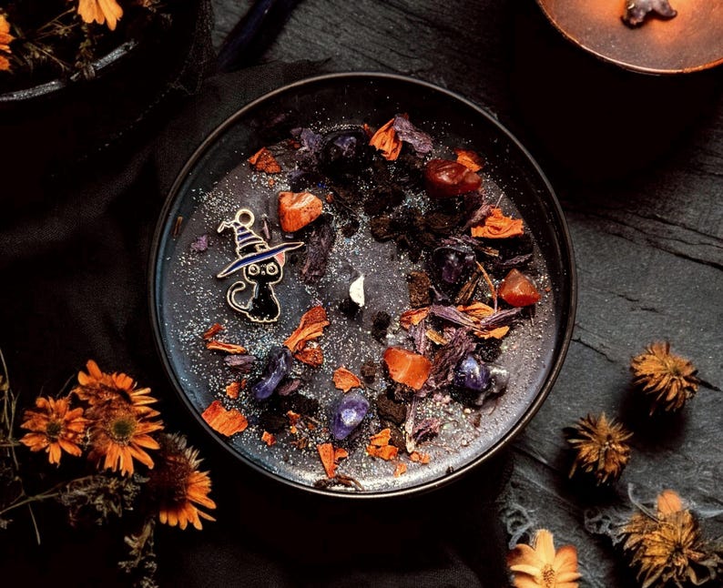 May include: A round black candle in a metal tin, topped with dark wax, silver glitter, dried orange flower petals, purple crystal chips, and a small metal charm of a black cat wearing a purple witch hat, centered around a single white wick.