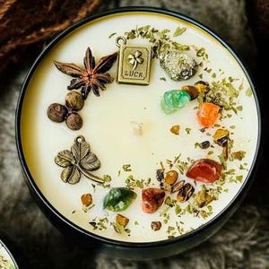 May include: A round black candle tin filled with cream-coloured wax, topped with a star anise, a metal charm shaped like a four-leaf clover, and a rectangular metal charm engraved with a clover and the word LUCK, alongside various colourful stones.