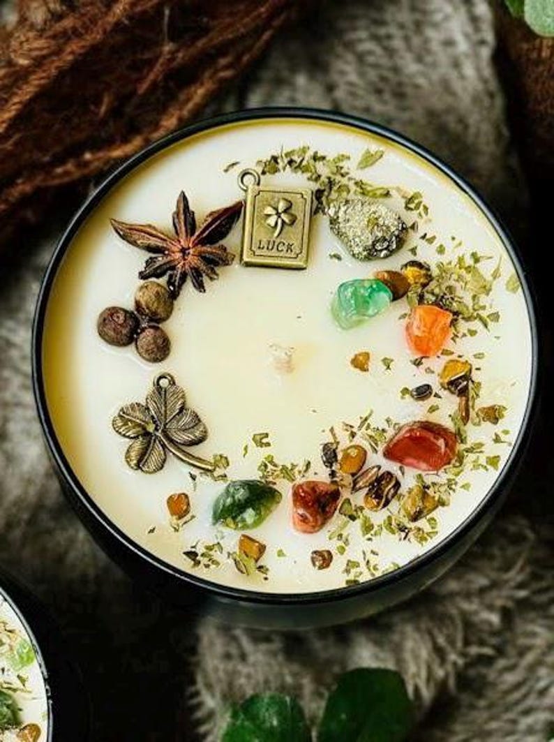 May include: A round black candle tin filled with cream-coloured wax, topped with a star anise, a metal charm shaped like a four-leaf clover, and a rectangular metal charm engraved with a clover and the word LUCK, alongside various colourful stones.