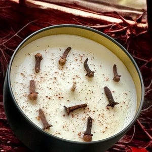 May include: A white candle with a black rim and cloves embedded in the wax. The candle is in a black tin container.