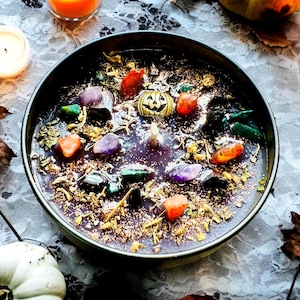 Peut inclure: Une bougie ronde dans un r&eacute;cipient sombre avec une surface de cire violette fonc&eacute;e incrust&eacute;e de diverses pierres pr&eacute;cieuses polies color&eacute;es, d'herbes s&eacute;ch&eacute;es, de paillettes dor&eacute;es et d'une petite breloque citrouille en m&eacute;tal dor&eacute;.