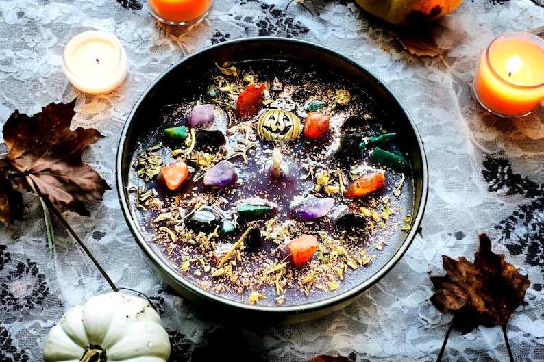 Peut inclure: Une bougie ronde dans un r&eacute;cipient sombre avec une surface de cire violette fonc&eacute;e incrust&eacute;e de diverses pierres pr&eacute;cieuses polies color&eacute;es, d'herbes s&eacute;ch&eacute;es, de paillettes dor&eacute;es et d'une petite breloque citrouille en m&eacute;tal dor&eacute;.