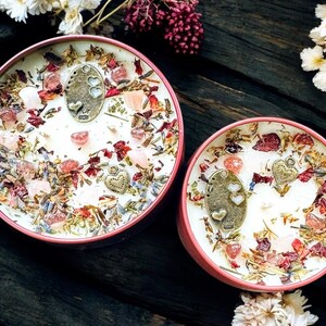 May include: Two round candles in pink metal tins, topped with white wax, dried flower petals, lavender sprigs, small pink crystals, and bronze-toned metal charms featuring heart cutouts and heart shapes embedded in the surface.