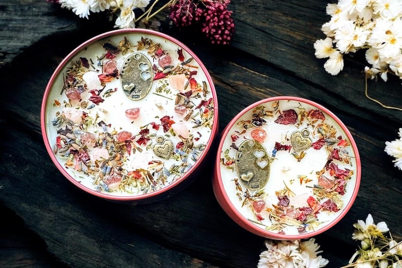 May include: Two round candles in pink metal tins, topped with white wax, dried flower petals, lavender sprigs, small pink crystals, and bronze-toned metal charms featuring heart cutouts and heart shapes embedded in the surface.