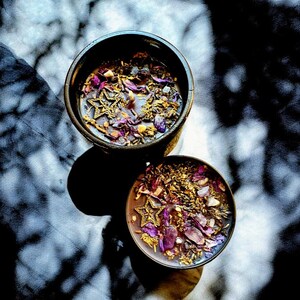 May include: Two black bowls filled with dried flowers, herbs, and gold glitter. The bowls are on a white surface with shadows from a window.