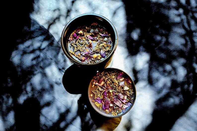 May include: Two black bowls filled with dried flowers, herbs, and gold glitter. The bowls are on a white surface with shadows from a window.