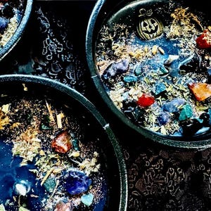 Peut inclure: Trois contenants &agrave; bougie circulaires noirs remplis de cire sombre, agr&eacute;ment&eacute;s de pierres pr&eacute;cieuses color&eacute;es, d'herbes s&eacute;ch&eacute;es et d'une breloque citrouille dor&eacute;e. La cire pr&eacute;sente un motif tourbillonnant bleu fonc&eacute; et noir avec des paillettes.