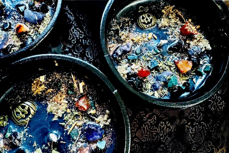 Peut inclure: Trois contenants &agrave; bougie circulaires noirs remplis de cire sombre, agr&eacute;ment&eacute;s de pierres pr&eacute;cieuses color&eacute;es, d'herbes s&eacute;ch&eacute;es et d'une breloque citrouille dor&eacute;e. La cire pr&eacute;sente un motif tourbillonnant bleu fonc&eacute; et noir avec des paillettes.