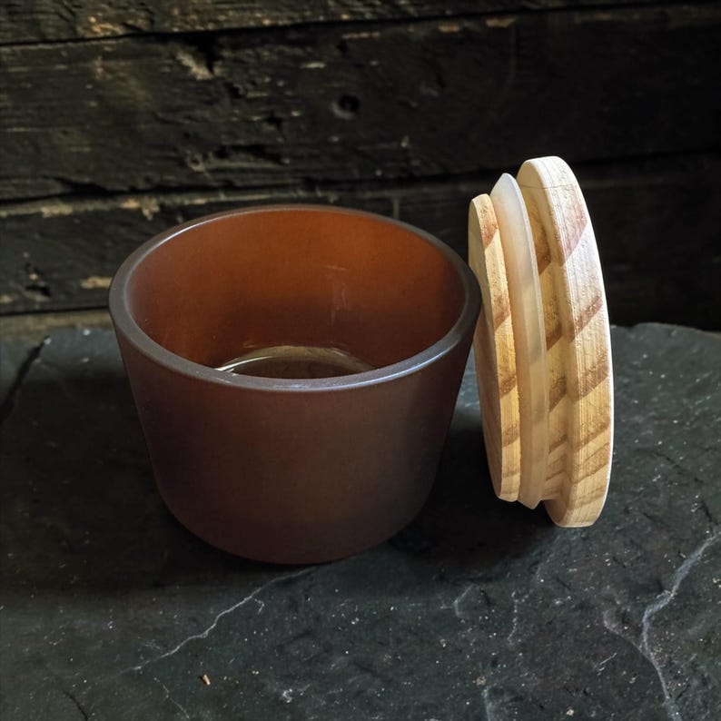 May include: A small, round, brown glass jar with a smooth, matte finish. The jar is open, and its circular wooden lid is placed upright next to it, showing a light-coloured wood grain pattern and a clear silicone seal ring on the underside.