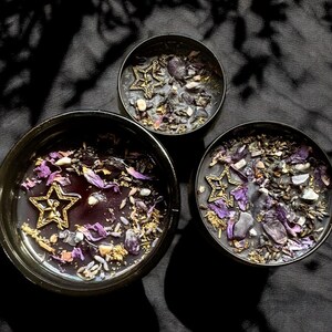 May include: Three black candle jars with purple and gold accents. Each jar contains a star-shaped charm and purple and gold crystals.