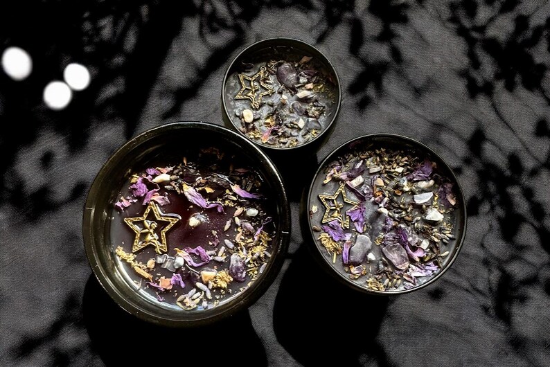 May include: Three black candle jars with purple and gold accents. Each jar contains a star-shaped charm and purple and gold crystals.
