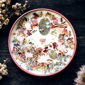 May include: A round candle in a pink metal tin, featuring a white wax surface embedded with dried flower petals, lavender sprigs, small pink crystal stones, and two metallic charms, one oval with heart cutouts and one heart-shaped pendant.