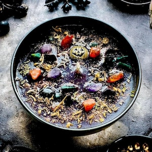 Peut inclure: Une bougie noire ronde dans un bol, garnie d'herbes s&eacute;ch&eacute;es, de paillettes, de diverses petites pierres pr&eacute;cieuses polies violettes, orange et vertes, et d'une breloque centrale en forme de citrouille dor&eacute;e avec un d&eacute;tail de cr&acirc;ne.