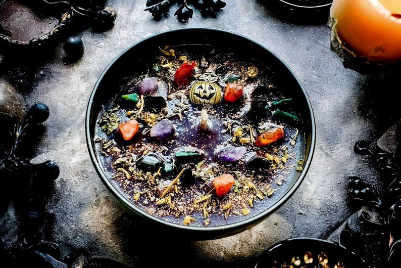 Peut inclure: Une bougie noire ronde dans un bol, garnie d'herbes s&eacute;ch&eacute;es, de paillettes, de diverses petites pierres pr&eacute;cieuses polies violettes, orange et vertes, et d'une breloque centrale en forme de citrouille dor&eacute;e avec un d&eacute;tail de cr&acirc;ne.